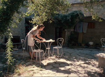Movie still from “La Belle Noiseuse” (1991), directed by Jacques Rivette – A woman standing at a table with chairs outside; Wide shot, High angle