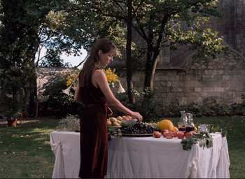 Movie still from “La Belle Noiseuse” (1991), directed by Jacques Rivette – A woman standing at a table with a lot of fruit; Wide shot, Over the shoulder angle