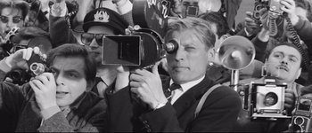 Movie still from “La Dolce Vita” (1960), directed by Federico Fellini – A man in a suit holding a video camera in front of a group of people; Close Up shot, Low angle