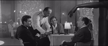 Movie still from “La Dolce Vita” (1960), directed by Federico Fellini – Two men standing at a table with a bottle of wine; Medium shot, Over the shoulder angle