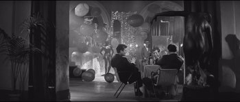 Movie still from “La Dolce Vita” (1960), directed by Federico Fellini – A black and white photo of a man sitting at a table in front of balloons; Wide shot, High angle