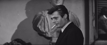 Movie still from “La Dolce Vita” (1960), directed by Federico Fellini – A black and white photo of a man smoking a cigarette in front of a statue; Close Up shot, Low angle