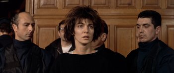 Movie still from “Nikita” (1990), directed by Luc Besson – A woman in a black shirt is standing in front of two other men; Close Up shot, Low angle