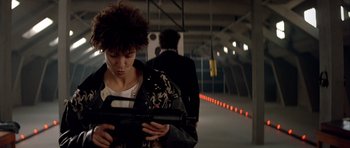 Movie still from “Nikita” (1990), directed by Luc Besson – A woman holding a gun while standing next to a wall; Medium shot, Over the shoulder angle