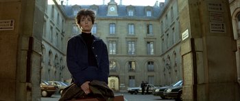 Movie still from “Nikita” (1990), directed by Luc Besson – A woman in a black jacket is standing in front of a building; Wide shot, Low angle