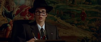 Movie still from “Nikita” (1990), directed by Luc Besson – A man in a suit and a hat is sitting at a desk; Close Up shot, Over the shoulder angle
