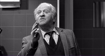 Movie still from “La haine” (1995), directed by Mathieu Kassovitz – An older man in a suit and tie is holding a cigarette; Medium shot, Over the shoulder angle