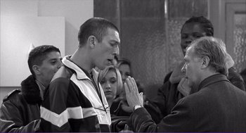 Movie still from “La haine” (1995), directed by Mathieu Kassovitz – A group of people gathered around a man with his hands clasped together; Medium shot, Over the shoulder angle