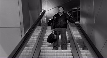 Movie still from “La haine” (1995), directed by Mathieu Kassovitz – A man is standing on an escalator holding a bag; Medium shot, Low angle