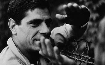 Movie still from “La Jetée” (1962), directed by Chris Marker – A black - and - white photo of a man holding a barbed wire fence; Close Up shot, Low angle