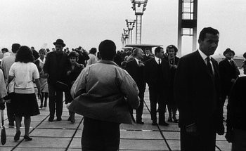 Movie still from “La Jetée” (1962), directed by Chris Marker – A group of people standing on top of a concrete walkway; Wide shot, Over the shoulder angle