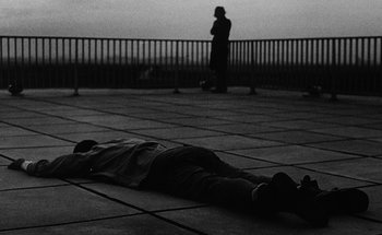 Movie still from “La Jetée” (1962), directed by Chris Marker – A man laying on the ground in front of a railing; Wide shot, High angle