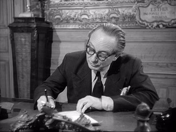 Movie still from “La Poison” (1951), directed by Sacha Guitry – An older man sitting at a table writing on a piece of paper; Medium shot, Over the shoulder angle