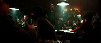 Movie still from “La Vie En Rose” (2007), directed by Olivier Dahan – A man standing in front of a group of people in a room; Medium shot, Low angle