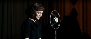 Movie still from “La Vie En Rose” (2007), directed by Olivier Dahan – A woman standing in front of a microphone in a dark room; Medium shot, Over the shoulder angle