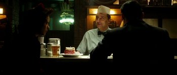 Movie still from “La Vie En Rose” (2007), directed by Olivier Dahan – A man sitting at a table in front of a plate of food; Medium shot, Over the shoulder angle