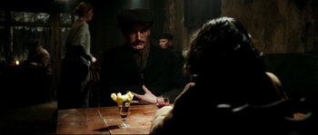 Movie still from “La Vie En Rose” (2007), directed by Olivier Dahan – A man sitting at a wooden table with a glass of wine; Medium shot, Over the shoulder angle