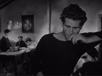 Movie still from “The Earth Trembles” (1948), directed by Luchino Visconti – A man smoking a cigarette in a room with other people; Medium shot, Over the shoulder angle