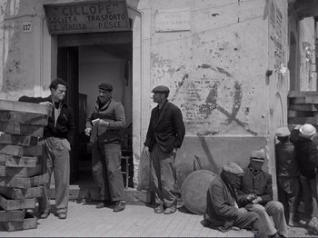 Movie still from “The Earth Trembles” (1948), directed by Luchino Visconti – A black and white photo of a group of men standing on the sidewalk; Wide shot, Low angle
