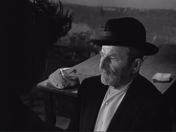 Movie still from “The Earth Trembles” (1948), directed by Luchino Visconti – An old man wearing a hat sitting next to a table; Medium shot, Over the shoulder angle