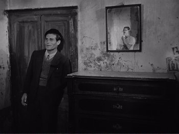 Movie still from “The Earth Trembles” (1948), directed by Luchino Visconti – A black and white photo of a man standing in front of a dresser; Medium shot, Over the shoulder angle