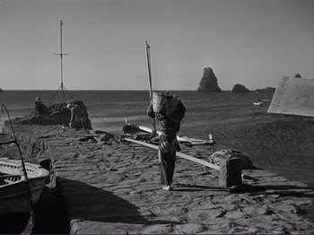Movie still from “The Earth Trembles” (1948), directed by Luchino Visconti – A man walking on the beach carrying a large bag; Extreme Wide shot, High angle