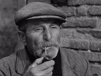 Movie still from “The Earth Trembles” (1948), directed by Luchino Visconti – An old man wearing a hat and smoking a pipe; Close Up shot, Low angle