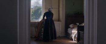Movie still from “Lady Macbeth” (2016), directed by William Oldroyd – An old woman standing in front of a window looking out at a bed; Wide shot, High angle