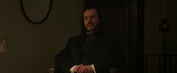 Movie still from “Lady Macbeth” (2016), directed by William Oldroyd – A man sitting in a chair wearing a suit and tie; Medium shot, Low angle