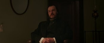 Movie still from “Lady Macbeth” (2016), directed by William Oldroyd – A man in a black suit sitting in a chair in front of a door; Medium shot, Over the shoulder angle