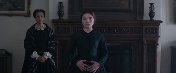Movie still from “Lady Macbeth” (2016), directed by William Oldroyd – A woman in a black dress standing in front of a fireplace; Medium shot, Over the shoulder angle