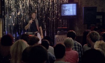 Movie still from “Ladybird Ladybird” (1994), directed by Ken Loach – A man standing in front of a microphone in front of a group of people; Wide shot, Over the shoulder angle