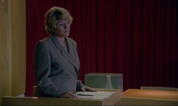 Movie still from “Ladybird Ladybird” (1994), directed by Ken Loach – A woman standing at a podium in front of a red curtain; Medium shot, Over the shoulder angle