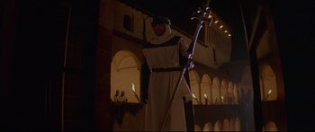 Movie still from “Ladyhawke” (1985), directed by Richard Donner – A man dressed as a monk holding a staff in front of a building at night; Wide shot, Low angle