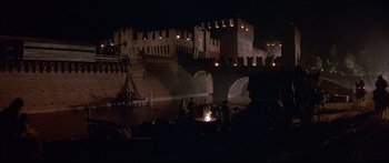 Movie still from “Ladyhawke” (1985), directed by Richard Donner – A group of people standing around a fire near a bridge; Extreme Wide shot, High angle