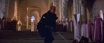 Movie still from “Ladyhawke” (1985), directed by Richard Donner – A man in a black coat holding a sword in front of a group of people; Medium shot, Over the shoulder angle