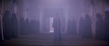 Movie still from “Ladyhawke” (1985), directed by Richard Donner – A woman standing in front of a group of people in a foggy room; Wide shot, High angle