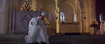 Movie still from “Ladyhawke” (1985), directed by Richard Donner – A man dressed as a priest sitting on a chair; Wide shot, High angle