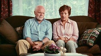 Movie still from “Lake Mungo” (2008), directed by Joel Anderson – An older man and woman sitting next to each other on a couch; Medium shot, High angle