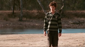 Movie still from “Lake Mungo” (2008), directed by Joel Anderson – A young man standing on a beach near a body of water; Medium shot, Over the shoulder angle