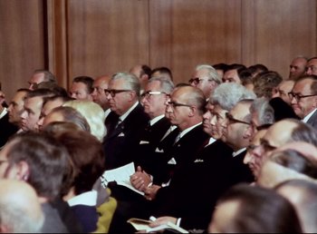 Movie still from “Land of Silence and Darkness” (1971), directed by Werner Herzog – A group of men in suits sitting next to each other in a room; Medium shot, High angle