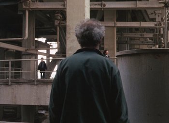 Movie still from “Landscape in the Mist” (1988), directed by Theodoros Angelopoulos – An older man is standing in an industrial setting; Wide shot, Over the shoulder angle