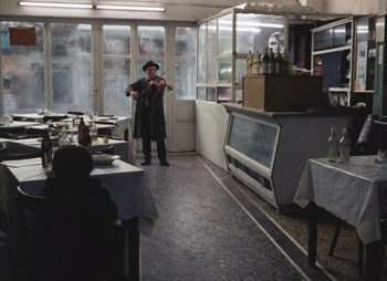 Movie still from “Landscape in the Mist” (1988), directed by Theodoros Angelopoulos – A man standing in a restaurant with a violin; Wide shot, High angle