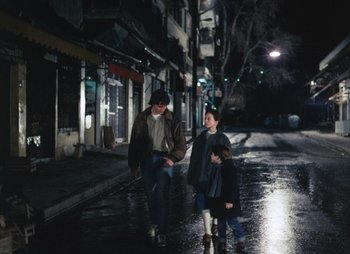 Movie still from “Landscape in the Mist” (1988), directed by Theodoros Angelopoulos – A man and two children walking down a street at night; Wide shot, High angle
