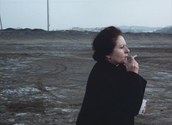 Movie still from “Landscape in the Mist” (1988), directed by Theodoros Angelopoulos – A woman standing in an open field smoking a cigarette; Medium shot, High angle