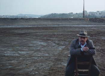 Movie still from “Landscape in the Mist” (1988), directed by Theodoros Angelopoulos – A man sitting on a chair in the middle of an empty field; Wide shot, High angle