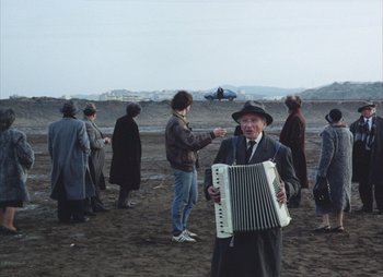 Movie still from “Landscape in the Mist” (1988), directed by Theodoros Angelopoulos – An old man holding an accordion in front of a group of people; Wide shot, High angle
