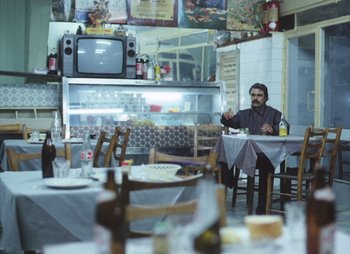 Movie still from “Landscape in the Mist” (1988), directed by Theodoros Angelopoulos – A man sitting at a table in front of a television; Wide shot, Low angle