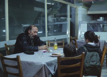 Movie still from “Landscape in the Mist” (1988), directed by Theodoros Angelopoulos – Three people are sitting at a table with food on it; Wide shot, Over the shoulder angle