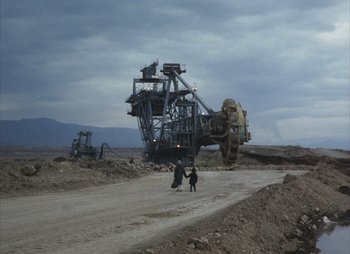 Movie still from “Landscape in the Mist” (1988), directed by Theodoros Angelopoulos – Two people are standing in front of a large machine; Extreme Wide shot, High angle
